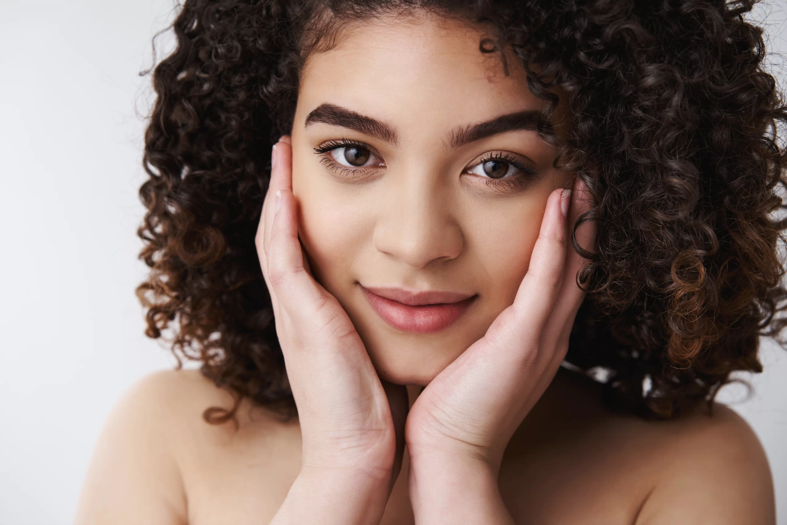 headshot-sensual-tender-attractive-naked-european-curly-haired-brunette-female-touch-pure-clean-skin-cheeks-look-camera-delighted-relaxed-cute-smile-take-care-flaws-imperfections-white-wall